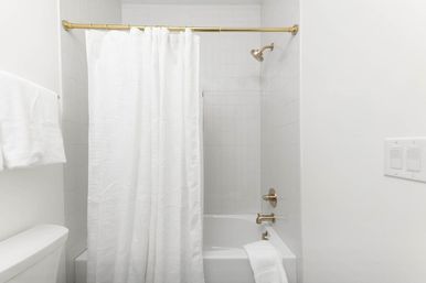 Minimal white bathroom with tub-shower combo, linen shower curtain on a brass rod, gold fixtures, white tile surround and folded towels
