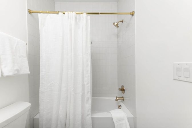 Minimal white bathroom with tub-shower combo, linen shower curtain on a brass rod, gold fixtures, white tile surround and folded towels