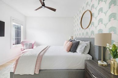 Bright, airy modern bedroom interior with a grey upholstered bed dressed in white linens and pink-and-grey pillows, mint-and-pink geometric wallpaper, round wooden mirror, wooden nightstand with lamp and gold vase, pink accent chair by a sunlit window and ceiling fan.
