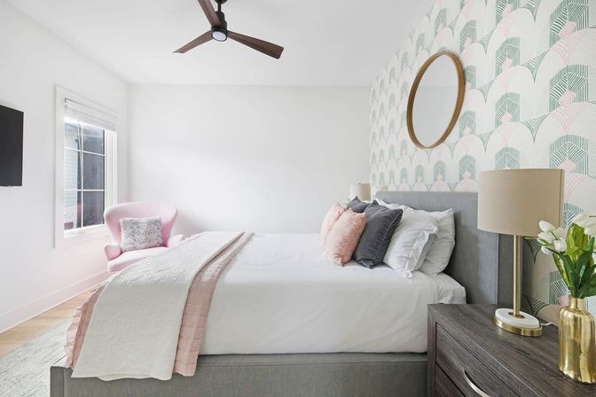 Bright, airy modern bedroom interior with a grey upholstered bed dressed in white linens and pink-and-grey pillows, mint-and-pink geometric wallpaper, round wooden mirror, wooden nightstand with lamp and gold vase, pink accent chair by a sunlit window and ceiling fan.