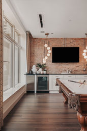 Stylish urban loft game room with exposed brick wall, wall-mounted TV, globe pendant lights, wet bar with mini fridge and decorative vases, large windows, hardwood floors, and a wooden pool table with cues.
