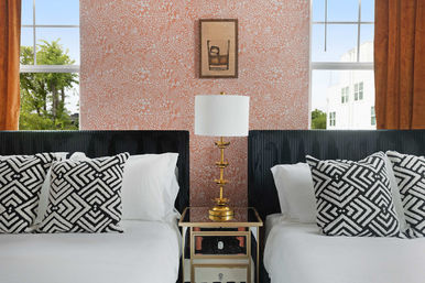 Cozy modern bedroom with two twin beds, black upholstered headboards, white linens and black-and-white geometric throw pillows, mirrored nightstand with a gold lamp, coral floral wallpaper and orange-curtain windows