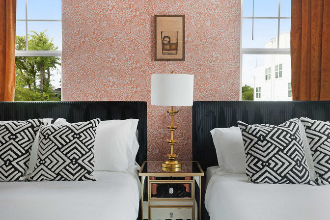 Cozy modern bedroom with two twin beds, black upholstered headboards, white linens and black-and-white geometric throw pillows, mirrored nightstand with a gold lamp, coral floral wallpaper and orange-curtain windows