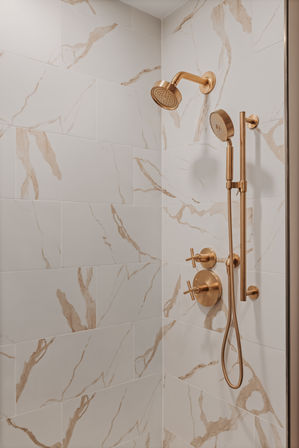 Corner bathroom shower with white marble-look tiles and brushed gold fixtures — wall-mounted showerhead, handheld wand, and cross-handle controls in a modern luxury spa-style design.