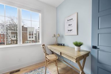 Bright modern home office with wooden desk and wicker chair by a large grid window overlooking urban rowhouses, light blue accent wall with framed 'learn live hope' print, desk lamp, potted plant and decorative letters.