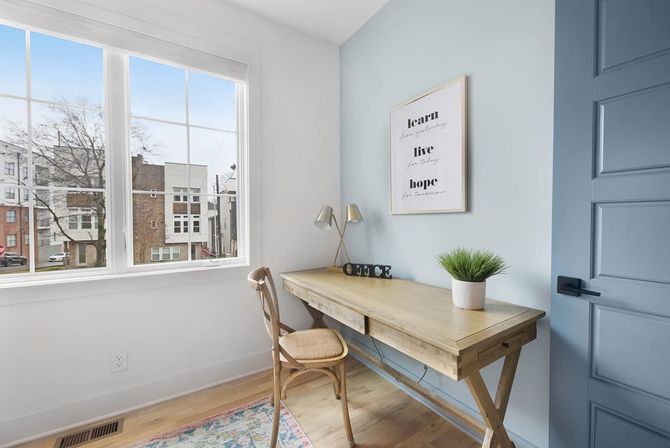 Bright modern home office with wooden desk and wicker chair by a large grid window overlooking urban rowhouses, light blue accent wall with framed 'learn live hope' print, desk lamp, potted plant and decorative letters.