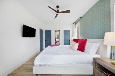 Bright modern bedroom interior with king bed in white linens, pink and red accent pillows, teal patterned accent wall, two blue doors, wall-mounted TV, wooden nightstand with lamp, ceiling fan, and hardwood floors.