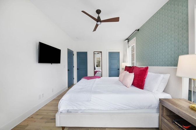 Bright modern bedroom interior with king bed in white linens, pink and red accent pillows, teal patterned accent wall, two blue doors, wall-mounted TV, wooden nightstand with lamp, ceiling fan, and hardwood floors.