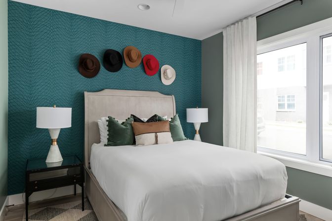 Sunlit modern bedroom with a teal textured accent wall, five hats hung above a neutral upholstered bed with white bedding and green velvet pillows, matching bedside lamps, black nightstand, and a large window with sheer curtains.