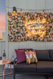 Rooftop patio lounge with a black sofa, colorful pillows, a pink and yellow flower wall and neon sign reading Smooth As Tennessee Whiskey, sunset sky and a side table with a bottle and glasses.
