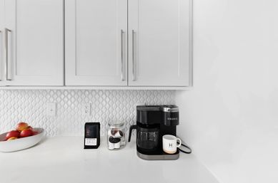 Minimal white kitchen coffee station on glossy countertop with a black single‑serve coffee maker and glass carafe, jar of coffee pods, bag of beans, white mug and bowl of red apples under white cabinets and textured tile backsplash.