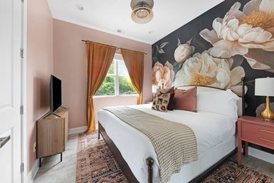 Cozy modern bedroom with a bold floral mural on a dark accent wall, white bed with textured throw and pink pillows, gold curtains at the window, wooden TV console, pink nightstand and patterned rug.