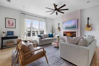 Sunlit modern living room with neutral sofas and leather mid-century chairs, large window, ceiling fan, wall-mounted TV over a fireplace, two guitars on the wall, hardwood floors and patterned rug.