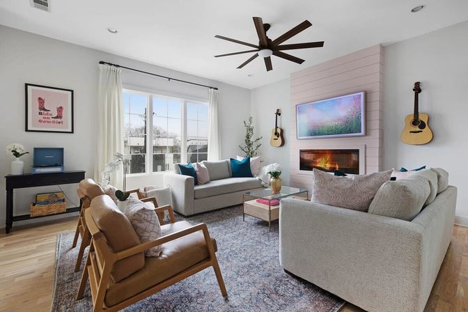 Sunlit modern living room with neutral sofas and leather mid-century chairs, large window, ceiling fan, wall-mounted TV over a fireplace, two guitars on the wall, hardwood floors and patterned rug.