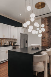 Modern urban kitchen with black waterfall island and cozy upholstered bar stools, sculptural cluster of globe pendant lights, white shaker cabinets with brass hardware, stainless refrigerator and exposed brick accent wall.