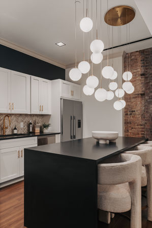 Modern urban kitchen with black waterfall island and cozy upholstered bar stools, sculptural cluster of globe pendant lights, white shaker cabinets with brass hardware, stainless refrigerator and exposed brick accent wall.