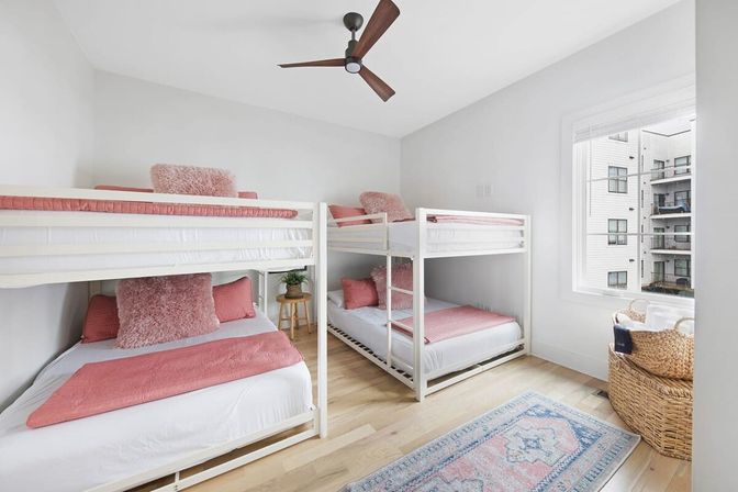 Bright, modern bunk bedroom with two white bunk beds dressed in pink linens and fluffy pillows, light wood floors, ceiling fan, woven basket by a sunlit window with apartment views, and a pastel area rug.