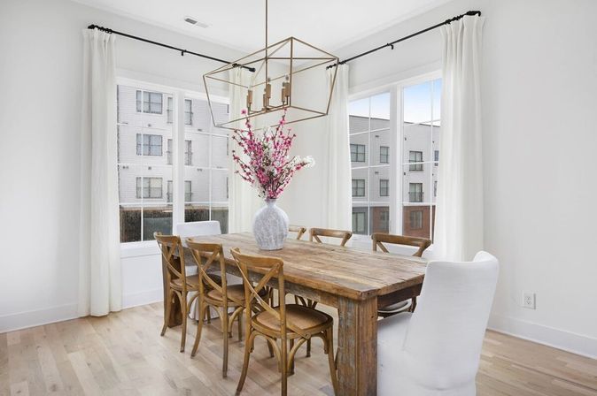 Bright modern dining room with rustic wooden farmhouse table, six chairs (wooden cross-back and white slipcovered ends), geometric gold chandelier, pink floral vase centerpiece, large windows with white curtains and an urban apartment view.