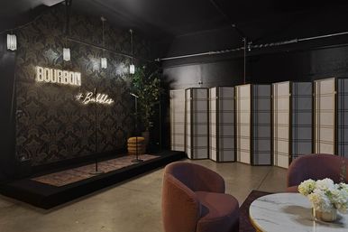 Industrial-chic event lounge with small stage, neon "Bourbon + Bubbles" sign, two microphones, patterned wallpaper, folding screens, cozy mauve chairs and a marble-top table with white flowers.