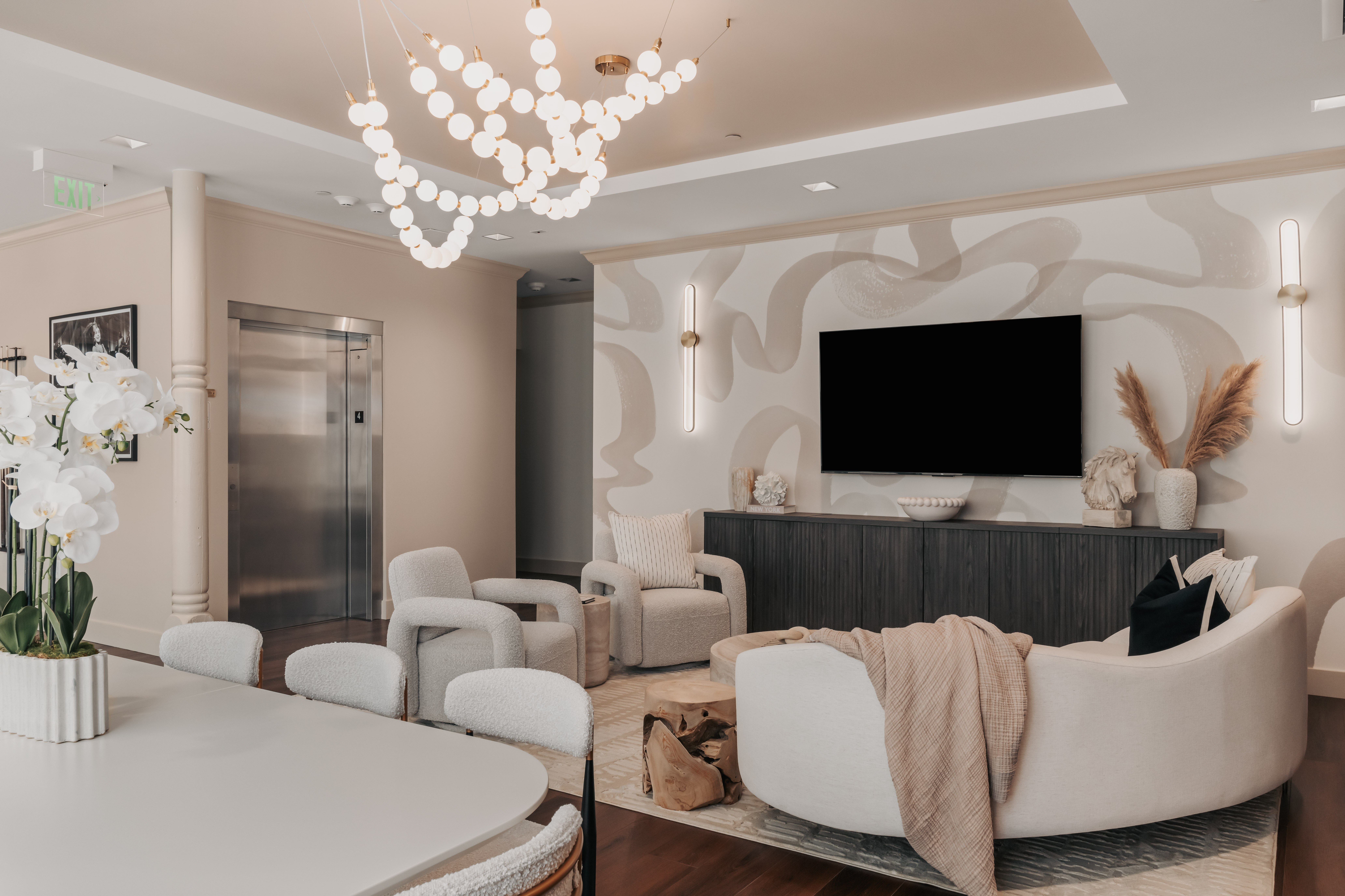 Neutral-toned modern lounge with curved sofa and boucle armchairs, wall-mounted TV, sculptural bubble chandelier, elevator doors, orchids and pampas grass accents.