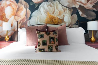 Stylish bedroom interior with oversized blush-and-white peony mural behind a crisp white bed, pink velvet pillows, framed-print accent cushion and gold bedside lamps on pink nightstands