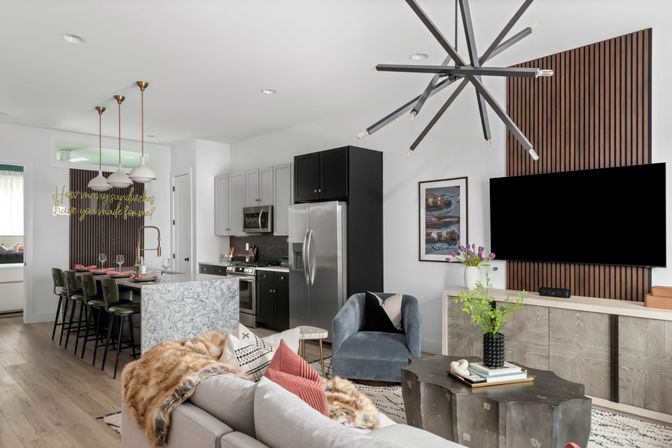 Modern open-concept living room and kitchen — marble island with green bar stools, stainless steel appliances, wood-slat TV wall, velvet armchair, sculptural chandelier, cozy sofa with faux-fur throw and indoor plants.