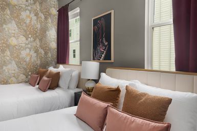 Cozy modern twin bedroom with two white beds, floral accent wallpaper, mauve curtains, pink and rust decorative pillows, bedside lamp and framed abstract art.
