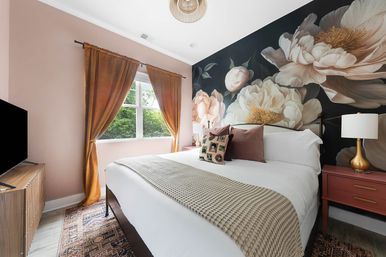 Cozy chic bedroom interior with a large dark floral mural behind a white bed, rust-gold curtains framing a sunlit window, pink nightstands with gold lamps, textured throw and patterned rug.