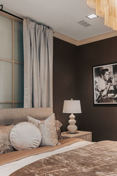 Cozy modern luxury master bedroom interior with plush beige bed, round and square decorative pillows, velvet duvet, wooden bedside table with sculpted white lamp, cream floor-to-ceiling curtains, dark accent wall and framed black-and-white portrait.