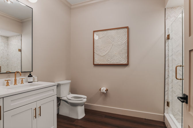 Modern neutral-toned home bathroom with white vanity, gold fixtures, glass shower, toilet, framed wall art and dark wood floor