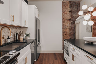 Chic modern urban kitchen with white cabinets, black countertops, brass faucet, stainless refrigerator, exposed brick accent wall, globe pendant lights, and warm hardwood floors.