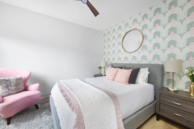 Chic modern bedroom with a gray upholstered bed dressed in white linens and blush-pink throws and pillows, mint-and-pink scallop wallpaper with a round gold mirror above the headboard, dark wood nightstands with lamps, and a pink accent armchair.