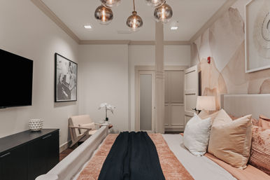 Cozy modern bedroom in blush and beige tones with a king bed, textured pillows and black throw, abstract watercolor accent wall, brass globe pendant lights, wall-mounted TV, armchair and orchid on a side table