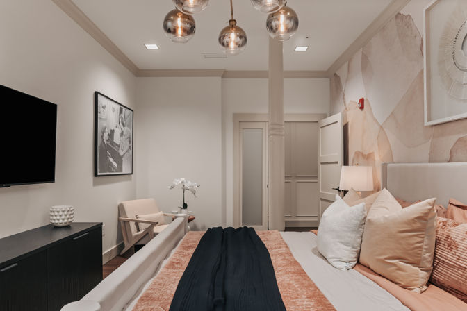 Cozy modern bedroom in blush and beige tones with a king bed, textured pillows and black throw, abstract watercolor accent wall, brass globe pendant lights, wall-mounted TV, armchair and orchid on a side table