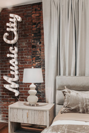 Cozy urban loft bedroom corner with exposed red brick wall, vertical glowing marquee sign, white sculptural table lamp on a light wood nightstand, beige upholstered headboard, neutral silk pillow and floor-to-ceiling curtains