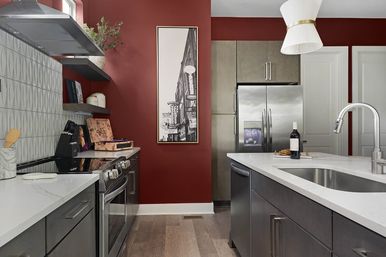 Contemporary urban kitchen with deep red accent wall, stainless steel refrigerator and range, white quartz countertops, center island with sink and wine bottle, geometric tile backsplash and light hardwood floors.