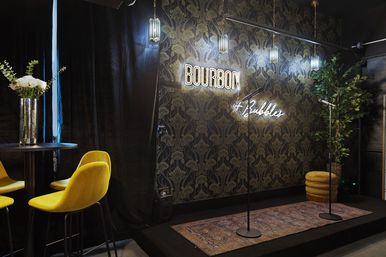 Intimate lounge stage with two microphone stands on a patterned rug, neon sign in mixed fonts on vintage wallpaper, pendant lights, yellow velvet bar stools and a small table with flowers.