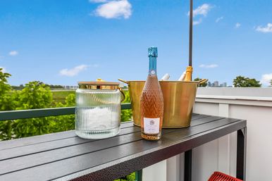 Rooftop balcony table with a sparkling rosé bottle, gold ice bucket holding extra bottles, and a ribbed glass jar, overlooking green treetops and a distant city skyline under a bright blue sky.
