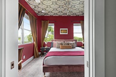 Cozy, stylish bedroom interior with burgundy walls and patterned ceiling, velvet upholstered bed with layered pillows and a red throw, gold curtains framing large windows with tree views, black nightstands with brass lamps and soft carpeted floor.