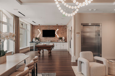 Stylish indoor lounge and game room with a wooden pool table, exposed brick accent wall and wall-mounted TV, large arched windows, globe pendant chandeliers, cozy boucle armchairs, a dining bar area, and stainless-steel elevator — modern apartment clubhouse vibe.