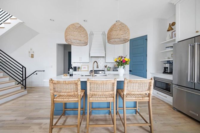 Sunlit modern coastal open-concept kitchen with blue island and white countertop, three woven rattan bar stools, matching pendant lights, stainless steel fridge, hardwood floors, and a vase of fresh flowers.