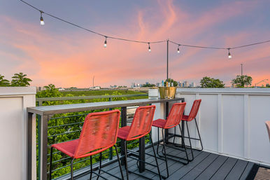 Rooftop balcony with four red woven bar stools at a metal high-top table, string lights overhead, trees below and a distant city skyline under a pink-orange sunset.