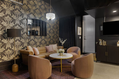 Cozy boutique lounge with tufted leather sofa and four blush swivel chairs around a round marble coffee table, black-and-gold floral wallpaper, crystal chandelier and gold accents.