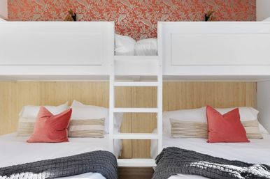 Bright coastal-style bunk bedroom with white built-in bunks and center ladder, two lower beds styled with coral throw pillows, striped lumbar cushions, gray knit blankets, bamboo panel headboard and coral floral wallpaper accent.