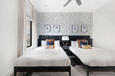 Modern urban apartment twin bedroom with two white-dressed beds, black upholstered headboards, geometric gray accent wall with framed art, central nightstand and lamp, blue and tan throw pillows, large window with curtains and ceiling fan.