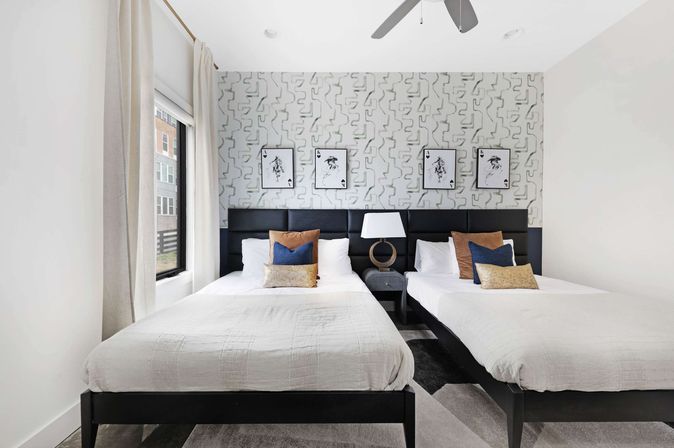 Modern urban apartment twin bedroom with two white-dressed beds, black upholstered headboards, geometric gray accent wall with framed art, central nightstand and lamp, blue and tan throw pillows, large window with curtains and ceiling fan.