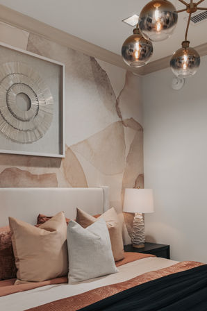 Cozy modern bedroom interior with beige upholstered headboard, layered terracotta and cream pillows, abstract watercolor accent wall, textured bedside lamp, and brass glass globe chandelier.