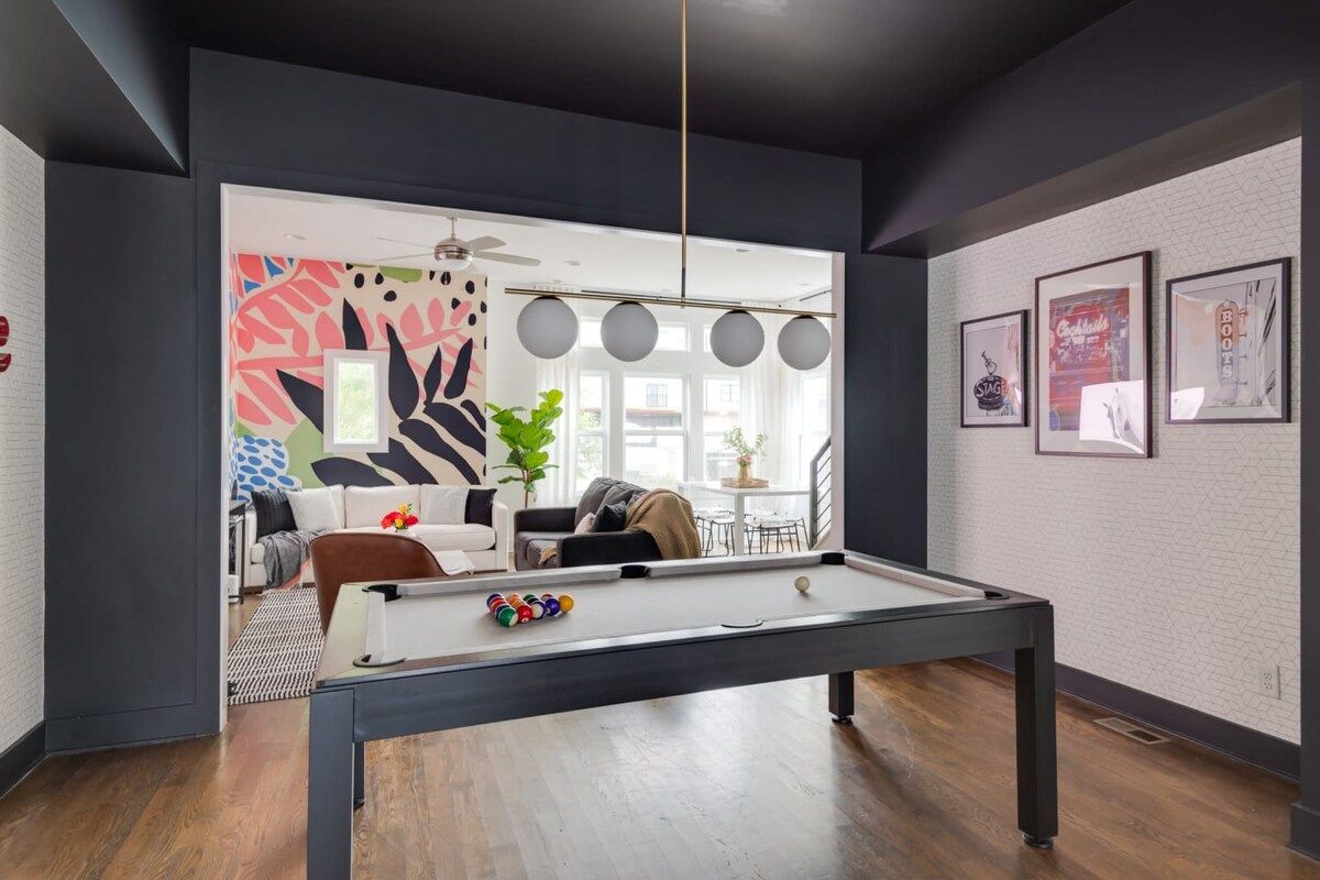Modern urban game room with a sleek black pool table on hardwood floors, open living area, bold tropical mural, globe pendant lights and sunlit windows.