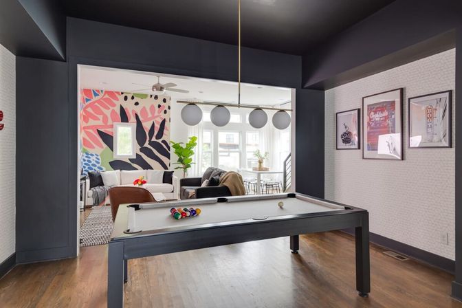 Modern urban game room with a sleek black pool table on hardwood floors, open living area, bold tropical mural, globe pendant lights and sunlit windows.