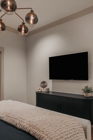 Cozy modern bedroom interior with wall-mounted flat-screen TV above a black ribbed media console, decorative vases and potted plant, plush beige textured throw on the bed, and a brass globe chandelier.